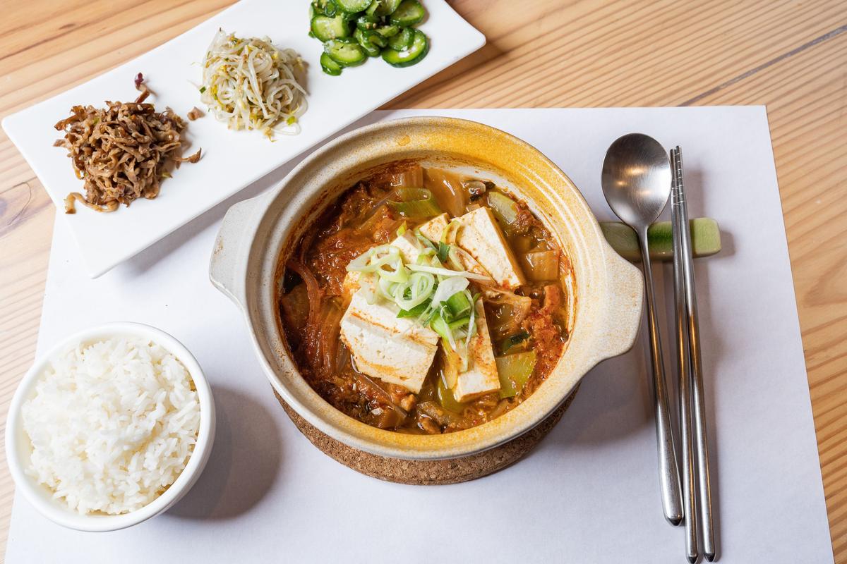 A single Korean meal for one: tofu stew in a pot with a bowl of rice, small side dishes and a spoon and chopsticks
