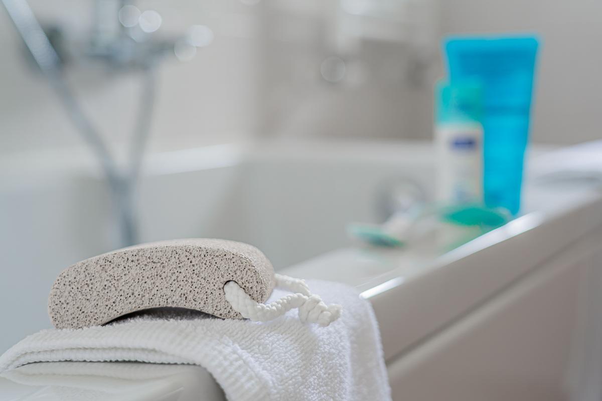 A pumice stone and a folded white towel on the edge of a bathtub, with toiletries behind