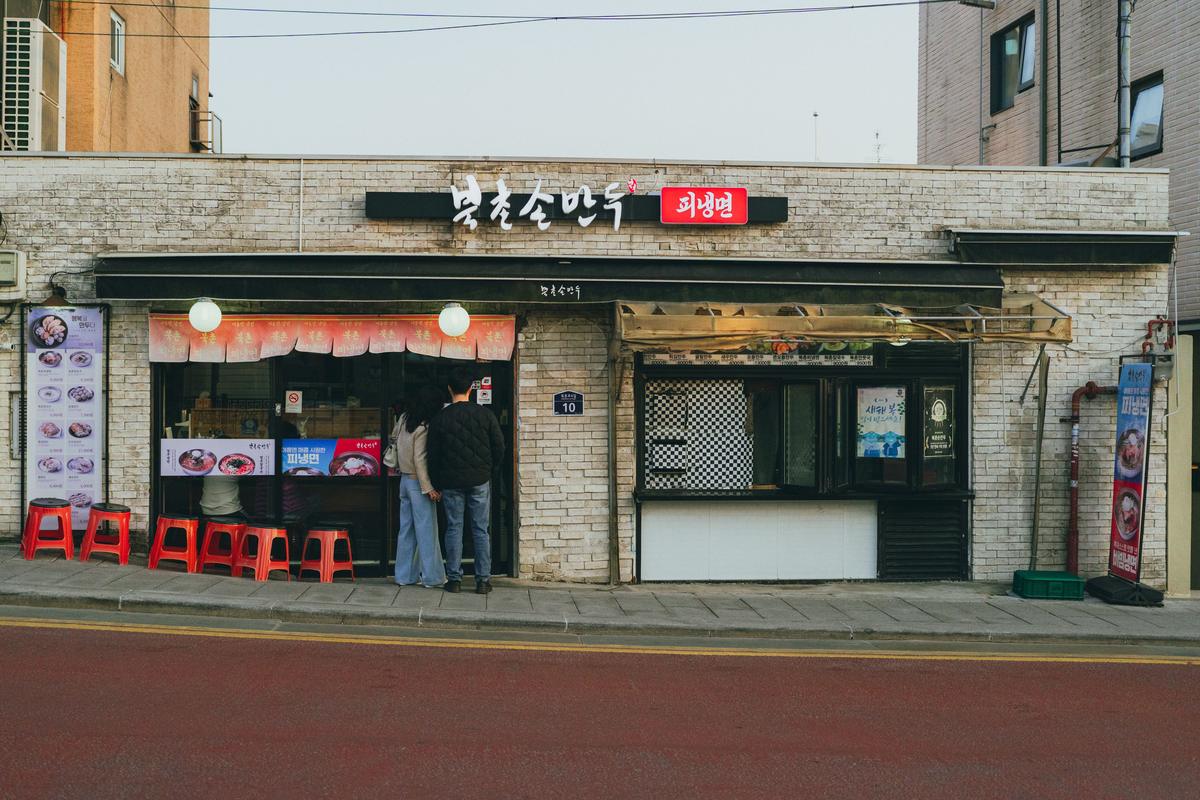 The front of a small Korean restaurant on a Seoul street, with red stools outside and a Korean sign above the door