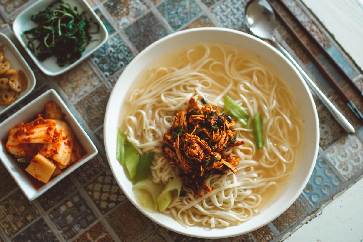 Overhead view of a bowl of Korean noodle soup with kimchi and small side dishes on a tiled table