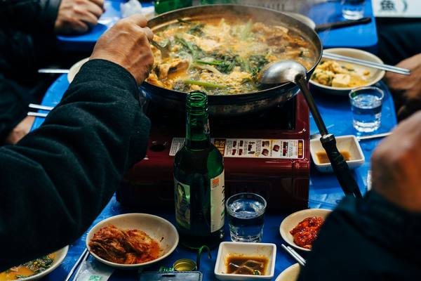 Diners in Seoul sharing a stew on a tabletop burner, with side dishes, soju and spoons in paper sleeves