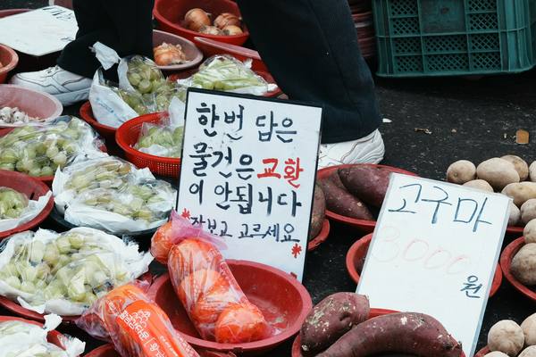 Vegetables at a street market with handwritten Korean price cards, one for sweet potatoes priced in won